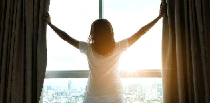 woman-opening-curtains-to-sunrise-