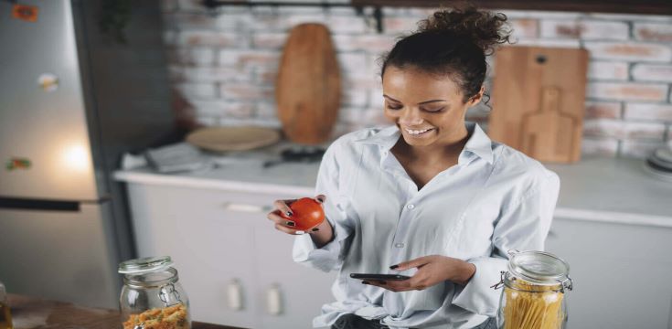 A-woman-is-smiling-at-her-phone-while-standing-in-the-kitchen-holding-an-apple