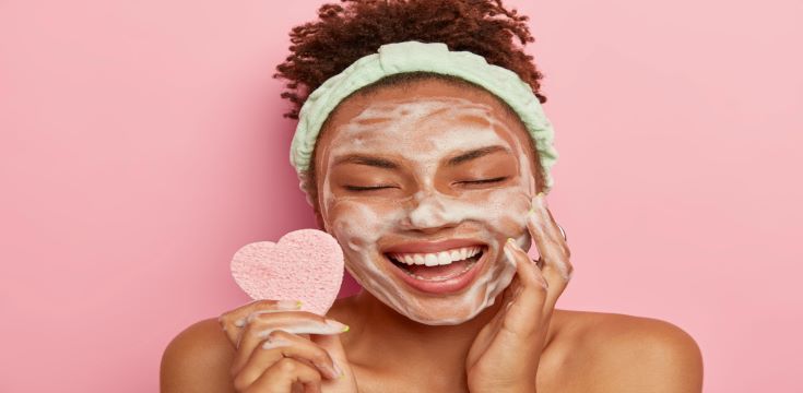 relaxed-joyful-dark-skinned-female-model-washes-face-with-soap-bubbles-enjoys-pampering-session-keeps-eyes-closed-from-pleasure-holds-cosmetic-sponge-cares-about-body-stands-naked-indoor