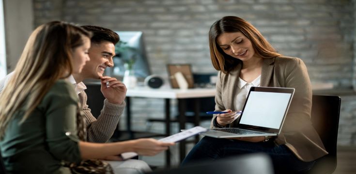 smiling-real-estate-agent-using-computer-while-having-meeting-with-young-couple-making-investment-plans