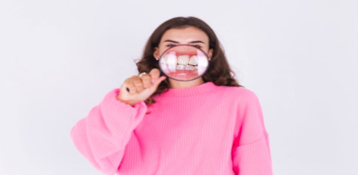 young-beautiful-woman-with-freckles-light-makeup-sweater-white-wall-with-magnifier-shows-white-teeth-perfect-smile