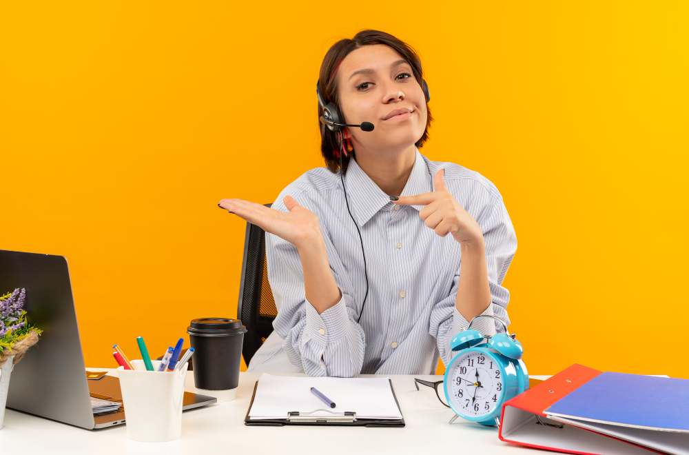 confident-young-call-center-girl-wearing-headset-sitting-desk-showing-empty-hand-pointing-it-isolated-orange
