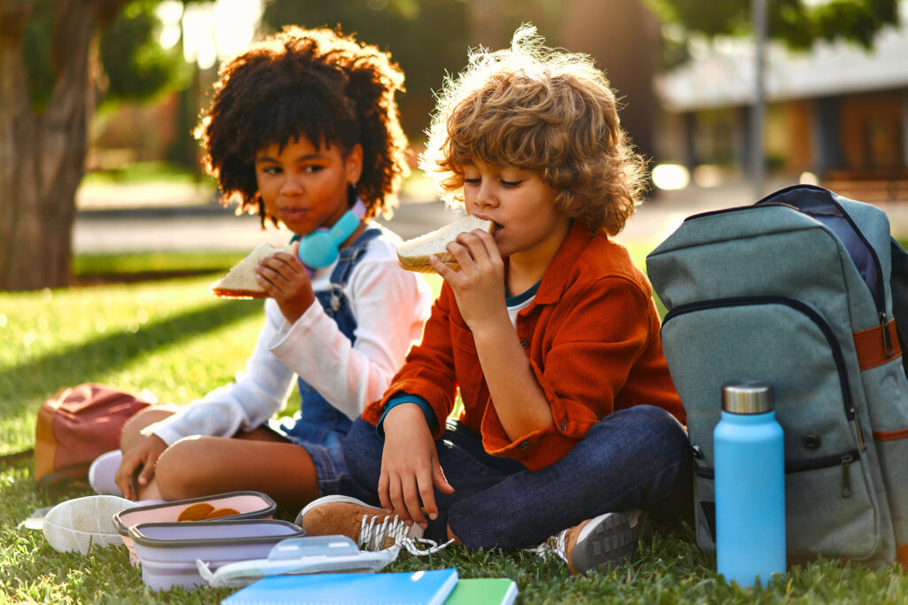 Charming schoolchildren after school resting sitting on the grass in the park. An African-American girl with a Caucasian boy took out lunchboxes with sandwiches from their backpack and eating.