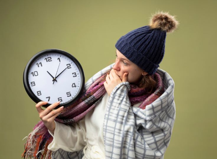 scared-young-ill-girl-wearing-white-robe-winter-hat-with-scarf-holding-looking-wall-clock-[15822]