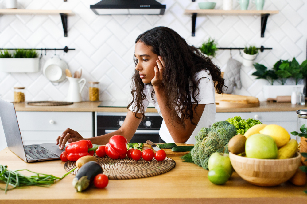 thoughtful-beautiful-mulatto-woman-is-looking-laptop-screen-modern-kitchen-table-full-vegetables-fruits-dressed-white-t-shirt