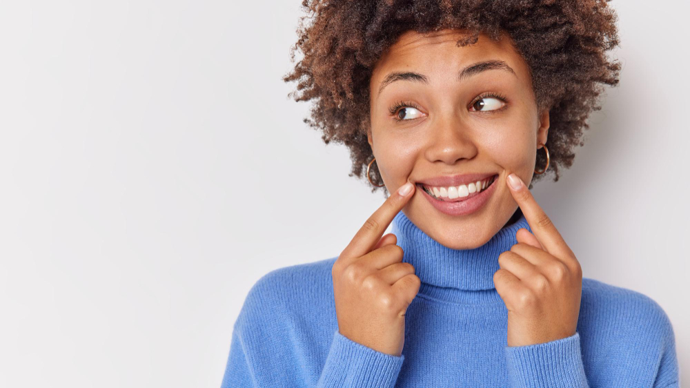 woman-smiles-broadly-points-her-perfect-even-teeth-indicates-corners-lips-looks-away-wears-casual-blue-jumper-isolated-white-with-blank-copy-space