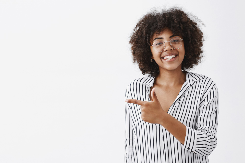 friendly-polite-good-looking-african-american-girl-glasses-with-curly-hair-striped-office-blouse-pointing-left-with-finger-gun-grinning-with-joy-giving-advice-where-go